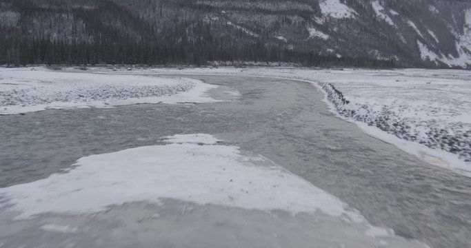 Aerial helicopter closup shot of partially frozen river with mountains in the distance, drone footage