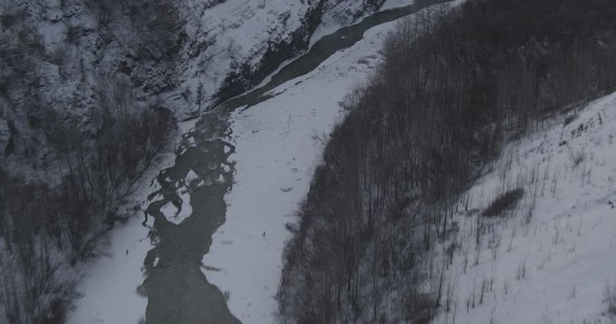 Aerial helicopter shot above winter valley with dark evergreens, over partially frozen lake towards mountains, drone footage