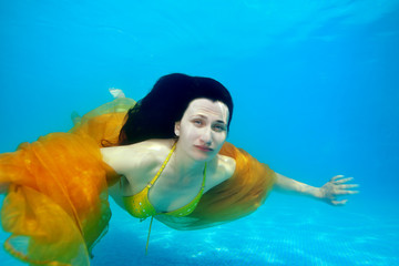 Obraz na płótnie Canvas A grown girl swims underwater near the bottom of the pool with her long hair down and a yellow cloth over her shoulders on a Sunny day. She looks at the camera. Surreal portrait