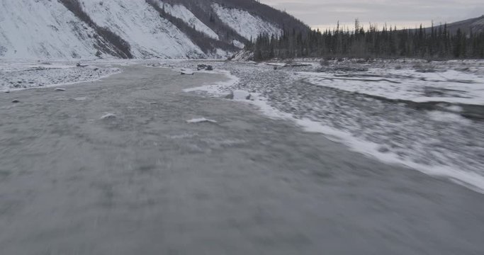 Aerial helicopter closup shot of partially frozen river, past snowy plateau with mountains in the distance, drone footage