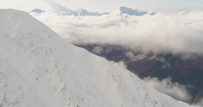Aerial helicopter shot, circling around large snow-capped mountain surrounded by clouds on bright and sunny day, drone footage
