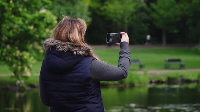 Girl stands with her back and takes a photo on her cell phone in Amsterdam park. Slow motion