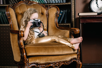 horizontal photo of happy beautiful little girl sitting in big golden cozy chair with photocamera. little cute vlogger.