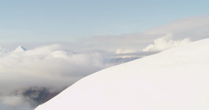 Aerial helicopter shot, circling around large snow-capped mountain in foreground to see mountain range surrounded by clouds in distance, drone footage