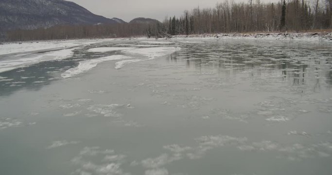  Aerial helicopter fly low over icy river, ice floes on water, trees, snow and tundra on banks mountains in distance, drone footage