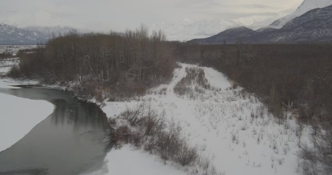  Aerial helicopter fly low through tree-covered riverside near frozen river in snowy mountain valley, drone footage