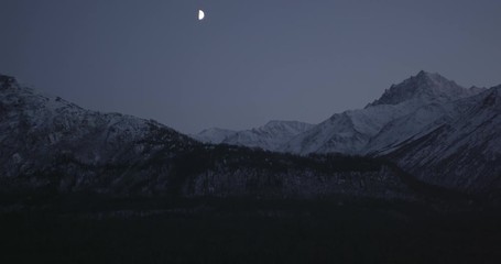 Aerial overhead helicopter shot, half moon gleams in between rocky and snowy Alaskan peaks, drone footage - Powered by Adobe