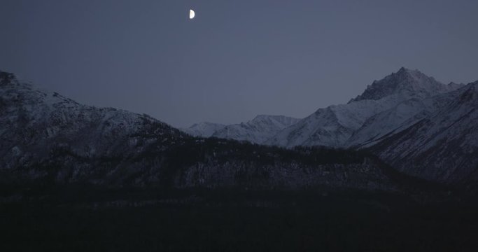  Aerial overhead helicopter shot, half moon gleams in between rocky and snowy Alaskan peaks, drone footage