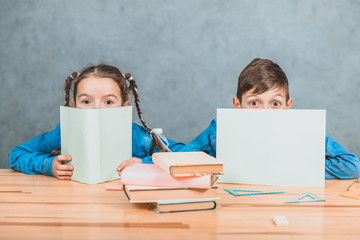 Curious little boy and girl hiding behind the sheets of papers, looking over, at the camera, conspiciuosly.