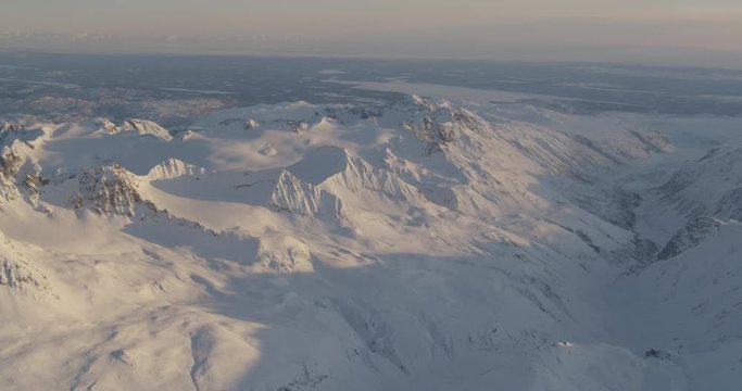  Aerial helicopter shot, panning across a vast, rocky, snowy Alaskan mountain range bathed in light during golden hour, drone footage, lens flare