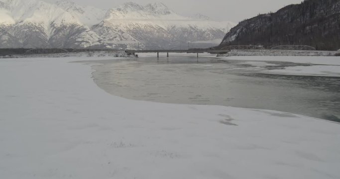  Aerial overhead helicopter shot, long push in on man standing in the center of snowy Alaskan bridge, he looks over the edges at the frozen water, car drives past in foreground, drone footage