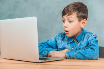 Cute little schoolboy sitting at the desk, surprised by something he has seen in his laptop.