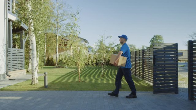 Delivery Man Holding Card Board Package Enters Through the Gates and Walks to the House and Knocks. Delivering Postal Parcel. In the Background Beautiful Suburban Neighbourhood. Following Side View