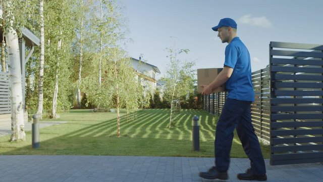 Delivery Man Holding Card Board Package Enters Through the Gates and Walks to the House and Knocks. Delivering Postal Parcel. In the Background Beautiful Suburban Neighbourhood. Following Side View