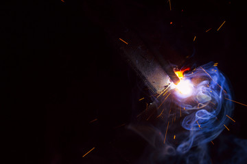 Obraz na płótnie Canvas Arc welding. Welding of two sheets of metal by electrode in inert gases. Type MMA. A bright flash of light and a sheaf of sparks in a cloud of smoke. Miniature Universe. Free space for inscriptions.