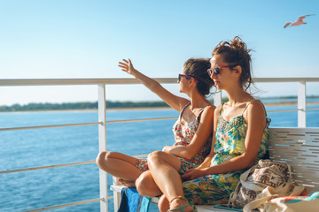 Two young women girl friends or sisters sitting on the bench on the deck of the ferry boat or ship sailing to the island tourist destination on summer vacation waving to the horizon in sunny day