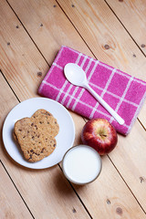 TOP VIEW OF BREAKFAST WITH COOKIES WITH CHOCOLATE CHIPS, MILK GLASS AND RED APPLE WITH KITCHEN CLOTH AND SPOON ON WOODEN TABLE. HEALTHY BREAKFAST