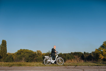 Fototapeta na wymiar Pensioner. An elderly man stand near an electric white bike on an asphalt road against the background of autumn nature. The concept of a happy old age and pension.