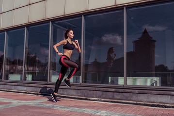 Naklejka na ściany i meble Beautiful athletic tanned athlete girl run summer city background glass windows, free space for fitness motivation text. Sportswear. Leggings. Top. Sneakers. Stylish fashionable slender woman.