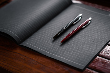 Two office pens in black and red located on a black notebook lying on a wooden table