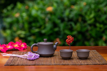 A small clay teapot with two cups for drinks standing on a mat surrounded by branches with flowers