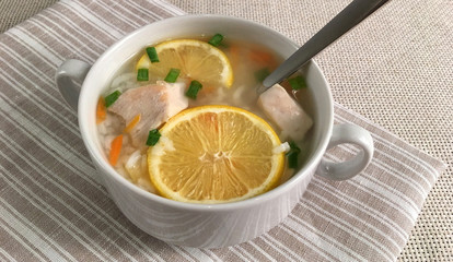Delicious chicken soup with lemon, rise, carrot and green onion. Top view. Soup with a spoon on a table. Rustic tablecloth. Striped serviette. Soup cup with chicken broth. b
