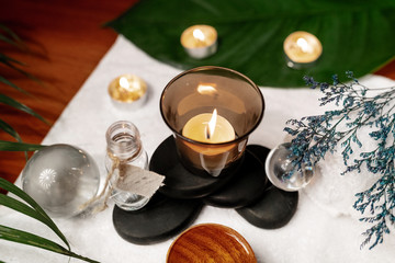 A lit candle standing on stones for a therapy stone with aromatic oil located on a terry towel next to which are transparent spheres, a twisted white terry towel and a sprig of lavender