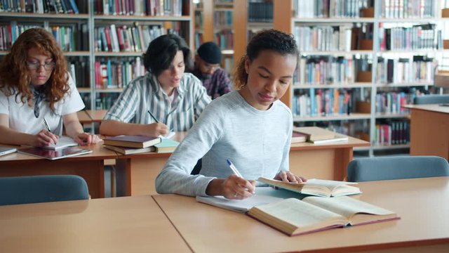 Slow motion of pretty mixed race girl studying in library with group of people writing reading then looking around relaxing. Education and smart youth concept.