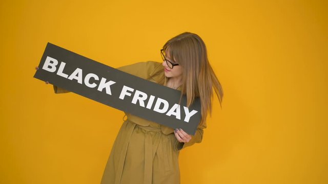 hipster european woman in glasses and fall trench coat with black friday sign isolated over yellow background