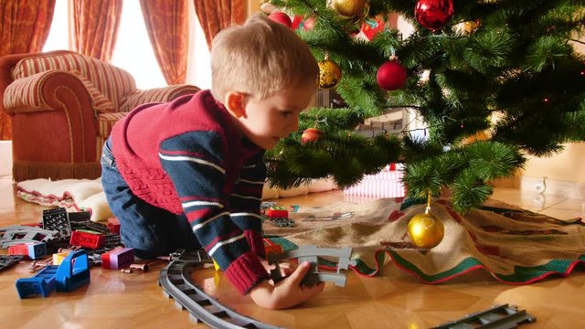 4k video of little boy recieved toy railraoad and train for Christmas from Santa Claus. Child building rails aroung Christmas tree in living room