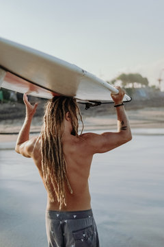 a young guy with dreadlocks carries a surfboard on his head, preparing to go riding the waves