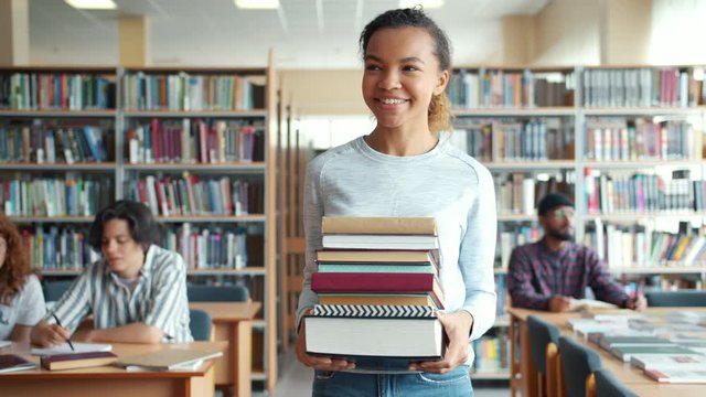 Pretty mixed race lady student is carrying books walking in library smiling while people studying reading at tables. Youth, education and literature concept.