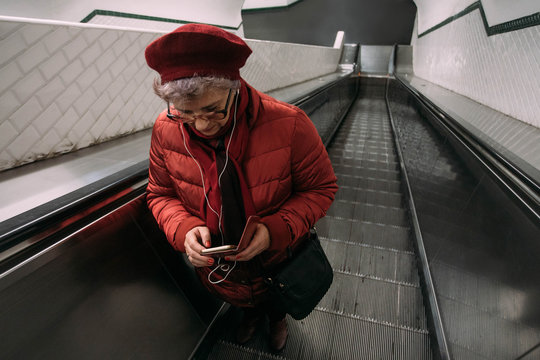 Senior woman standing at the escalator in Paris subway station, using smartphone with headphone