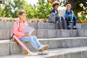 Portrait of girl sitting on stairs with skateboard and smiling at camera while reading a book with her classmates in the background