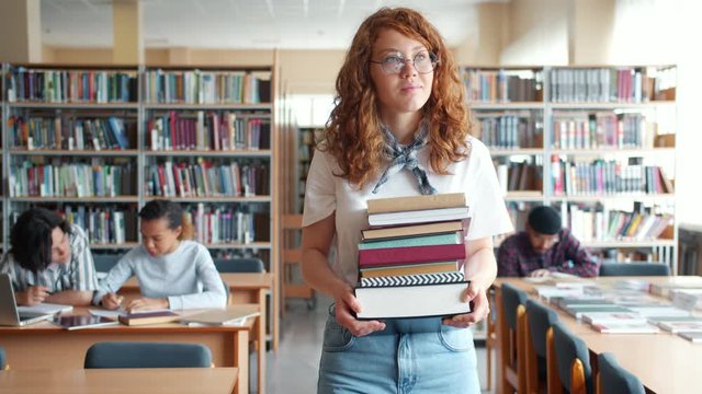 Slow motion of young female librarian walking in library with books smiling looking around at desks and students. People, occupation and workplace concept.