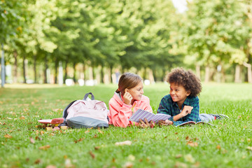 Two school children lying together on green grass with books and talking to each other during reading