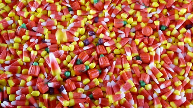 A large pile of assorted Halloween candy corn and pumpkins are shown. Pieces of the candy are seen dropping into the pile.