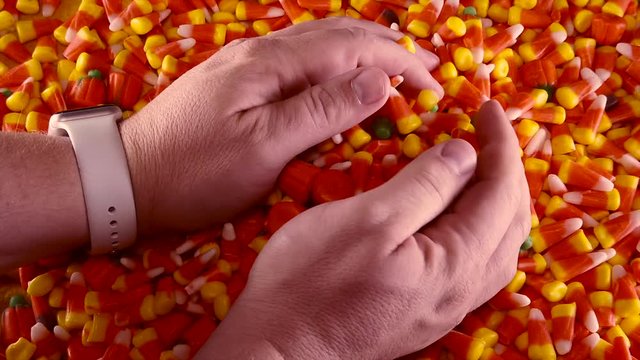Two hands are seen grabbing into a large pile of Halloween candy corn and pumpkins.