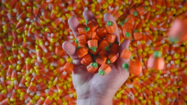 A large pile of Halloween candy corn is shown. A hand is seen above the candy corn with candy pumpkins dropping on top of it.