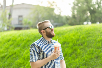 Side view of a stylish young man with a beard holding a milkshake and admiring the city views walking in the park on a warm summer day. The concept of rest and relaxation.