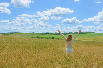 Young free woman with blowing hair outstretched arms, spreading hands with joy and inspiration facing the field, enjoying life. Girl in wheat field. Colorful field landscape