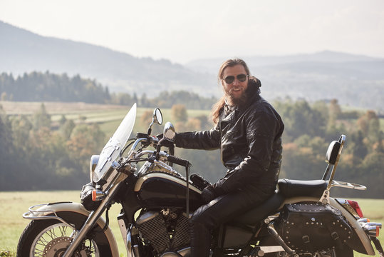 Handsome bearded motorcyclist with long hair in black leather jacket and sunglasses sitting on cruiser motorcycle, on blurred background of green peaceful rural landscape and light foggy sky.