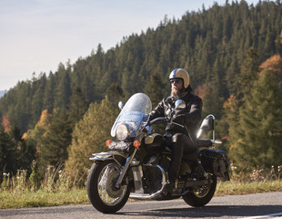 Handsome bearded biker in black leather jacket and sunglasses sitting on cruiser motorcycle on country roadside, on blurred background of foggy green hills covered with dense spruce forest. - obrazy, fototapety, plakaty