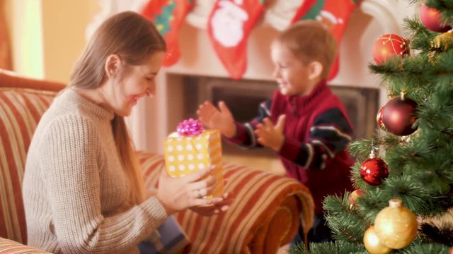 4k video of happy smiling little boy giving Christmas present box with ribbon to his mother next to Christmas tree. Child receiving gifts and presents from Santa Claus on winter holidays and