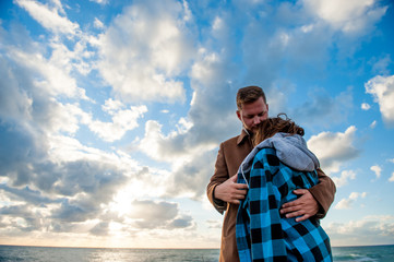 love and care concept young couple man in autumn coat hugging his shivering woman in plaid shirt and hoodie on blue sky with clouds and sea background