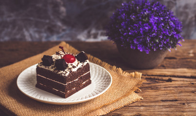 Black forest cake decorate with cherry and cake brownies In a white plate, place are on sackcloth and have false heather In pots small, all place are on wooden table.