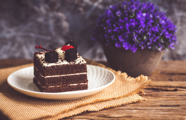 Black forest cake decorate with cherry and cake brownies In a white plate, place are on sackcloth and have false heather In pots small, all place are on wooden table.