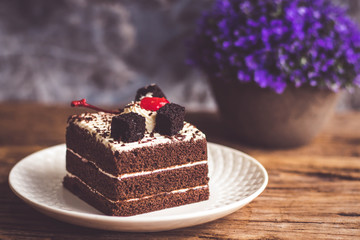 Black forest cake decorate with cherry and cake brownies In a white plate,and have false heather In pots small, all place are on wooden table.
