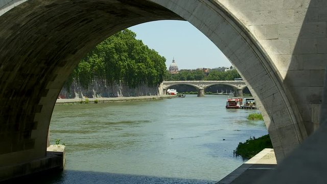 Wide low angle daytime still shot of Tiber River through Ponter Umberto bridge arch, stationary ship in water, and Cathedral dome against blue sky background, Rome, Italy