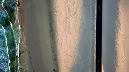Aerial above view of yellow tropical sandy beach with black long way road and car traveling - ocean blue waves and shore - sunset time with long beautiful shadow - concept of summer vacation